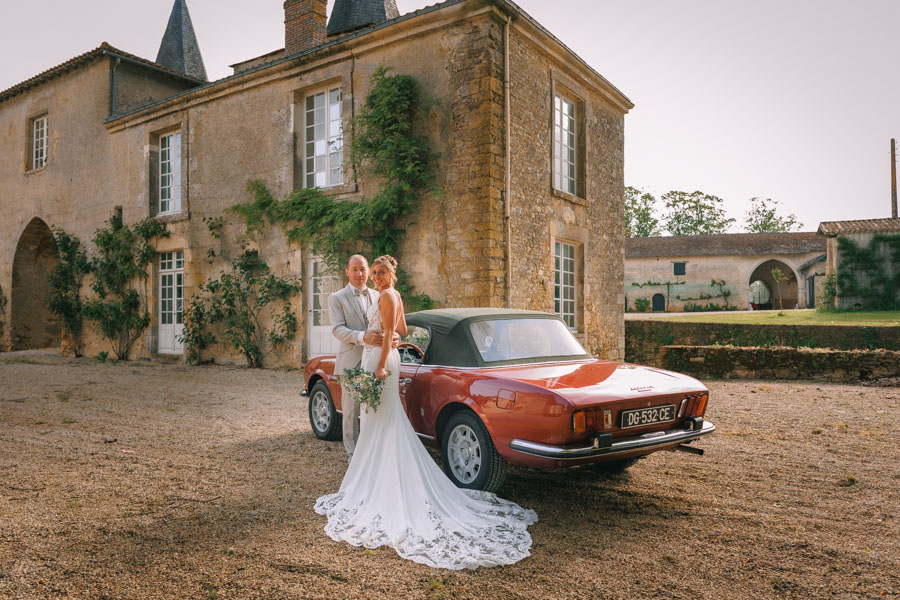 Un couple de marié posent devant un château et un voiture pour les photos de leur photographe de mariage en Vendée